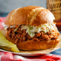 A pulled pork sandwich topped with coleslaw sitting beside a dill pickle.