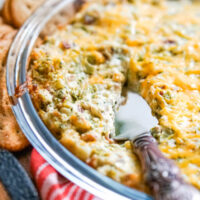 A pie plate of cheesy bacon and broccoli dip with a bronze server.