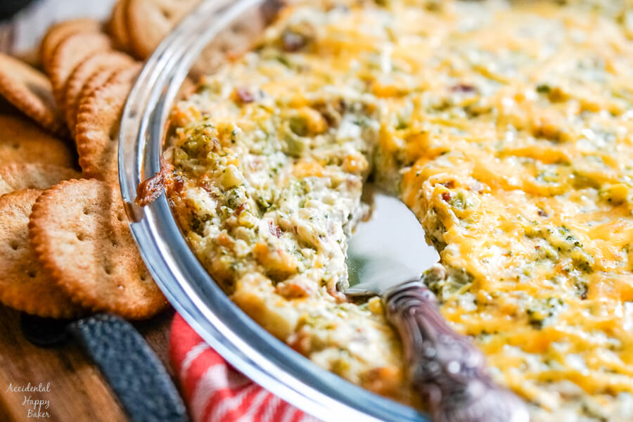 A pie plate of cheesy bacon and broccoli dip with a bronze server. 